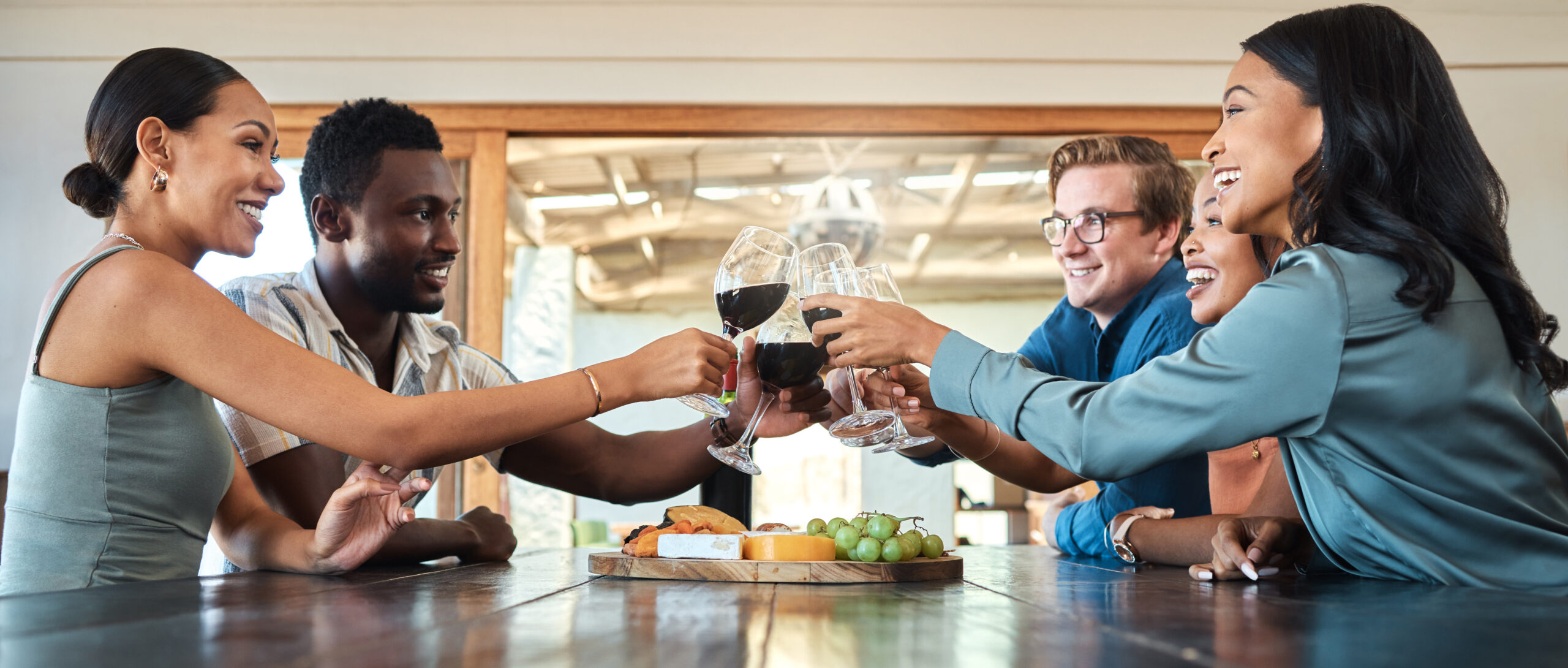 Group of people sitting around a table, toasting with glasses of red wine, with a cheese and fruit platter in the center