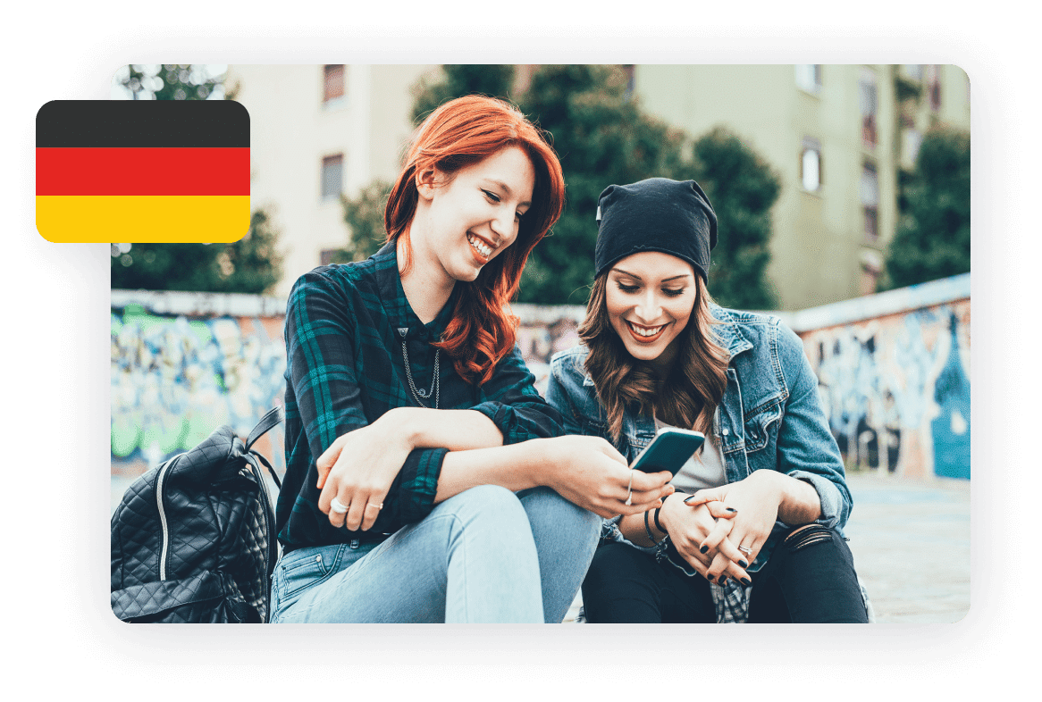 Two young women sitting outdoors, smiling and looking at a smartphone. One has red hair and the other has brown hair and is wearing a beanie. The German flag is displayed in the upper left corner.